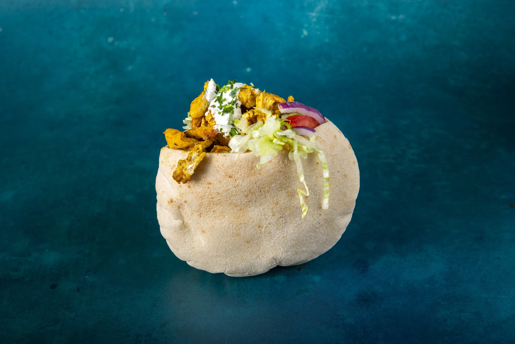 A pita filled with chicken, tzatziki, shredded iceberg lettuce, tomato and red onion, photographed against a deep teal background