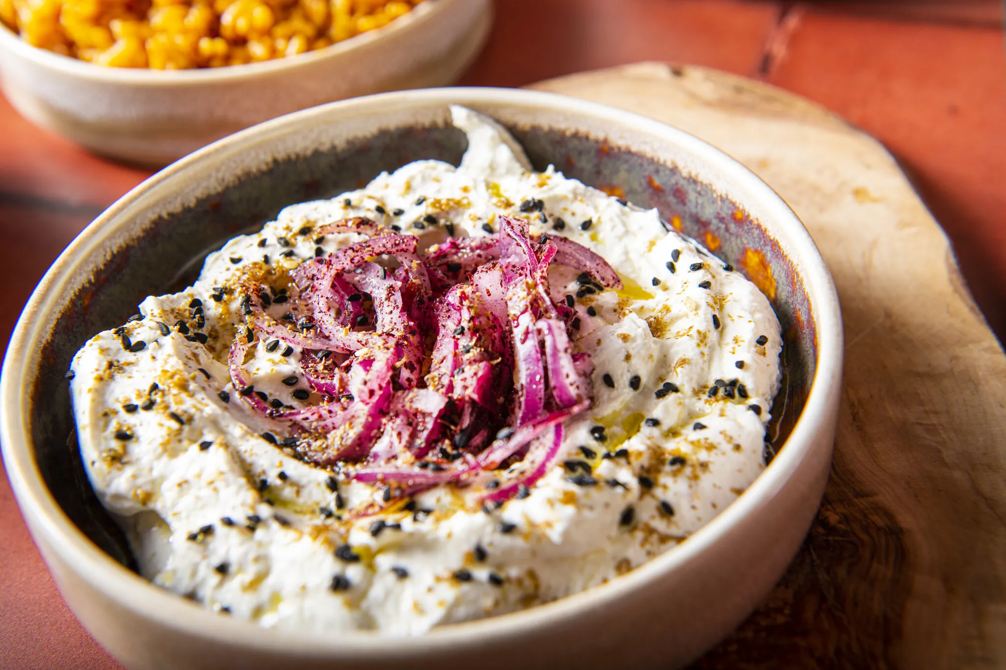 A bowl of labneh topped with sumac onions, black sesame seeds, za'atar and olive oil, on a terracotta table at ZHUG
