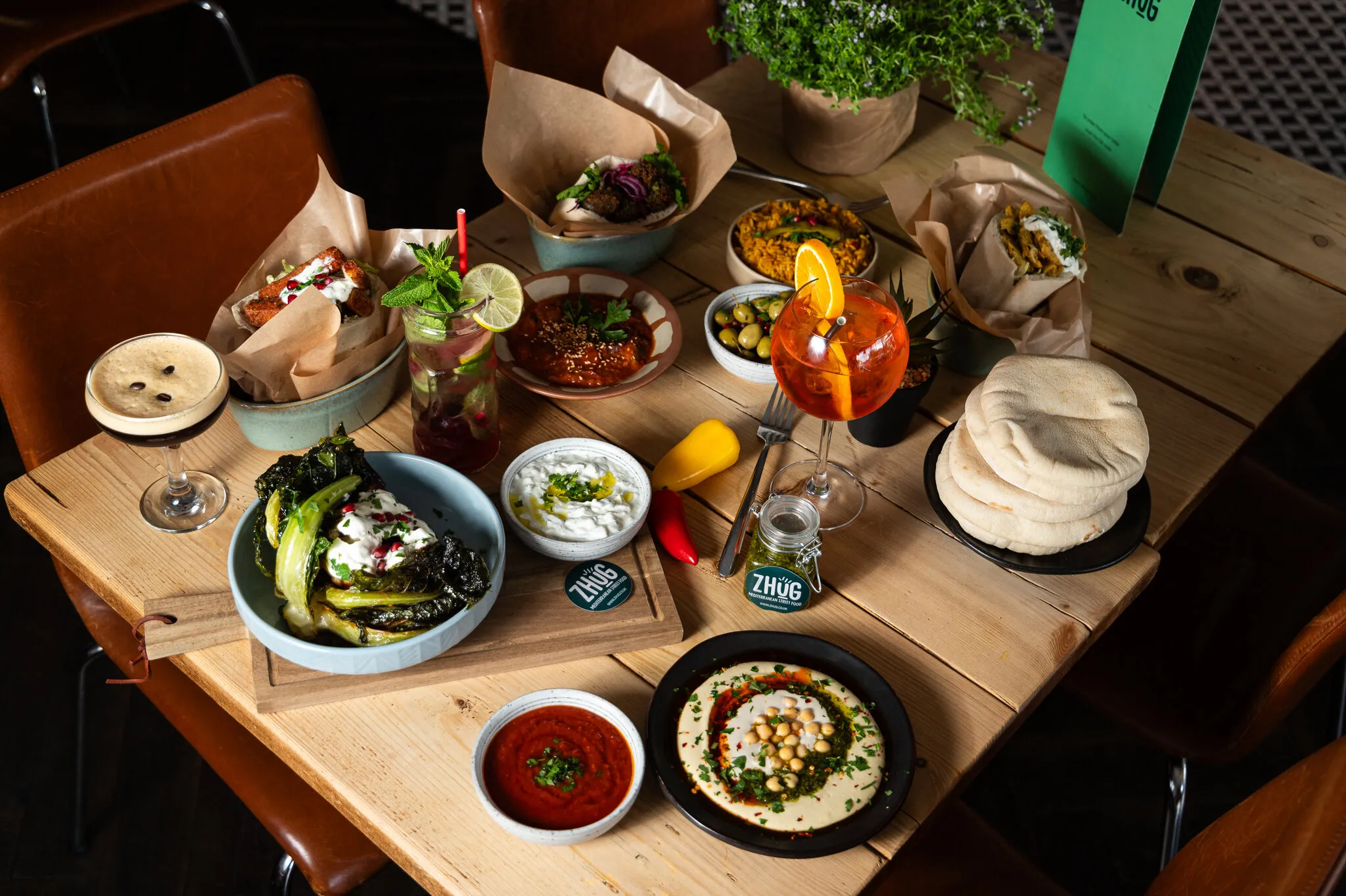 A wooden table at ZHUG laid with hummus, a stack of warm pita, olives, rice, pitas wrapped in paper and three cocktails